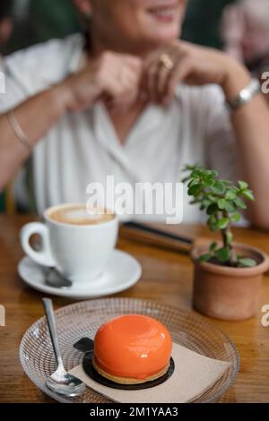 Bella donna caucasica di mezza età seduta a un tavolo in un bar e sorridente verso gli amici mentre si gusta una tazza calda di caffè al latte e dessert Foto Stock