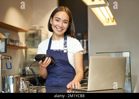 Barista asiatico sorridente, ragazza con terminale carta, macchina per il pagamento e computer portatile, in piedi in caffetteria, elaborazione pagamento per ordine caffè Foto Stock