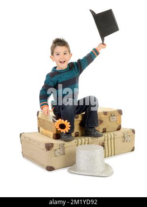 Ragazzo isolato in jeans e pullover a righe seduto su alcune vecchie valigie gioca in un cappello. Foto Stock