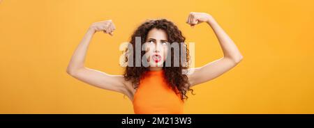 Donna sensazione potente e forte alzare le mani con pugni clenched facendo faccia intensa che si sta allenando in palestra mostrando muscoli e bicipiti guardando Foto Stock