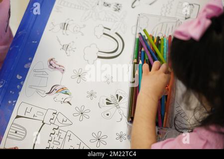 Un bambino siede e colora un gigantesco motivo di carta da colorare. Il bambino tiene in mano una matita. Un set di matite colorate su una scatola. Attività di disegno. Foto Stock