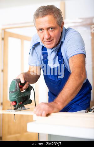 Concentrated male builder in uniform using a jigsaw machine on wooden plank at the apartment Foto Stock