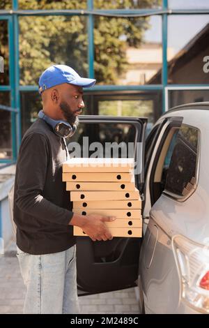 Corriere afroamericano che trasporta la pizza all'ufficio dalla vista laterale dell'automobile, mucchio delle scatole di ritegno del deliveryman. Addetto al servizio di consegna del pranzo che trasporta fastfood, in piedi vicino all'edificio aziendale all'aperto Foto Stock