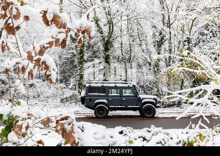 Un Land Rover Defender originale di colore nero è parcheggiato sotto alberi pieni di neve.alberi e foglie circondano questo splendido motore di trazione a quattro ruote nero Foto Stock