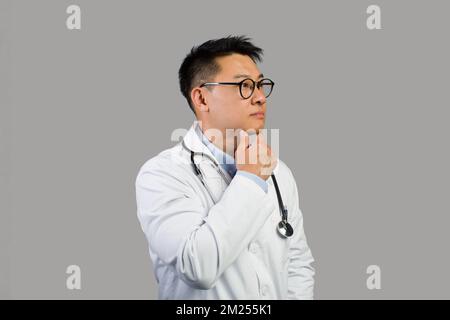 Pensivo sicuro uomo asiatico medio-anziano medico in camice bianco, occhiali con stetoscopio, pensa, guarda lo spazio libero Foto Stock