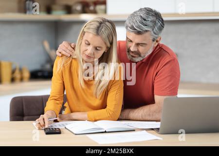 Mature spouses sitting at kitchen desk in front of computer Foto Stock