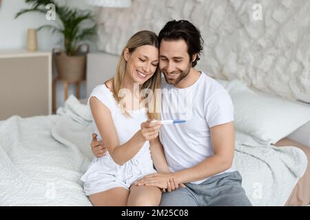 Happy husband and wife rejoicing positive pregnancy test, sitting on bed in bedroom interior, copy space Stock Photo