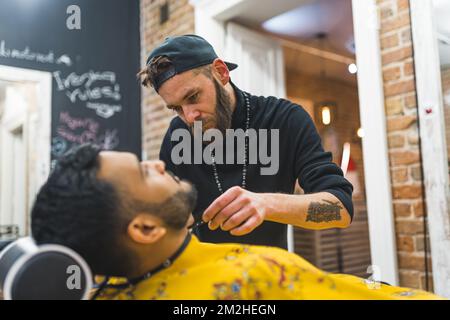 Professional bearded barber trims the beard of his client. Blurred Indian adult man in the foreground. Stylish hairstylist at barbershop. High quality photo Foto Stock