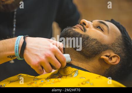 Primo piano della rifinitura professionale della barba. Barbiere irriconoscibile che utilizza attrezzature per parrucchieri per rifinire la barba del suo cliente indiano. Foto di alta qualità Foto Stock