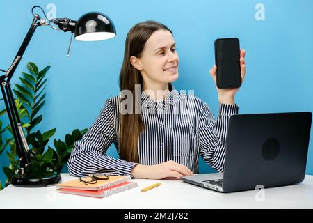 Felice donna di successo del lavoro del dipendente in camicia seduta lavoro alla scrivania bianca ufficio con laptop che tiene il telefono cellulare schermo vuoto area di lavoro, Foto Stock