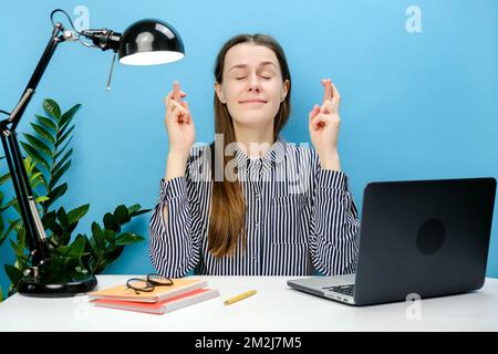 Donna dipendente di successo cute sedersi al lavoro in ufficio con PC laptop attendere momento speciale tenere le dita incrociate facendo desiderio, in posa isolato su blu Foto Stock