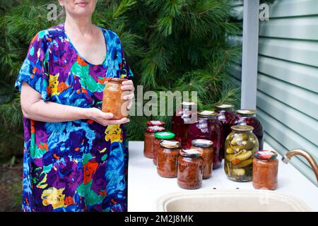 La vecchia donna tiene in mano un vasetto di marmellata fatta in casa. Modello anziano in abito blu sullo sfondo di cibi in scatola artigianali con sottaceti preservati Foto Stock