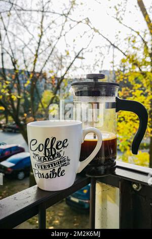 Frenchpress con caffè nero fresco accanto a una piccola tazza di caffè bianco in piedi su una ringhiera metallica di un piccolo balcone Foto Stock