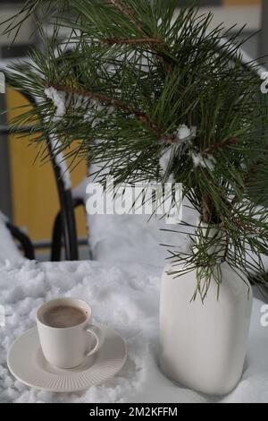 foto un ramo di abeti in un vaso bianco si trova accanto a una tazza di caffè con caffè su un tavolo coperto di neve Foto Stock