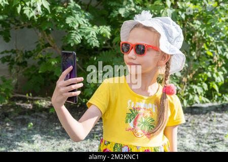 Felice ragazza sorridente bambino che cerca nel telefono cellulare all'aperto in estate. Una ragazza allegra vestita in giallo, un cappello bianco e occhiali da sole regge uno smar Foto Stock