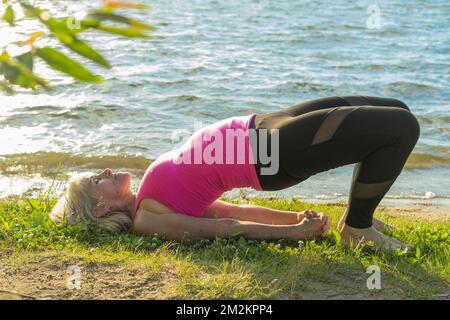 Una donna in abbigliamento sportivo in piedi in un ponte yoga posa sulla riva del lago. Urdhva Dhanurasana I. Meditazione. Yoga nel parco. Sport estivi Foto Stock