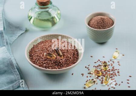 Semi di lino marroni o semi di lino e semi di lino macinati o schiacciati in una piccola ciotola e capsule di gelatina con olio omega su sfondo azzurro. Foto Stock