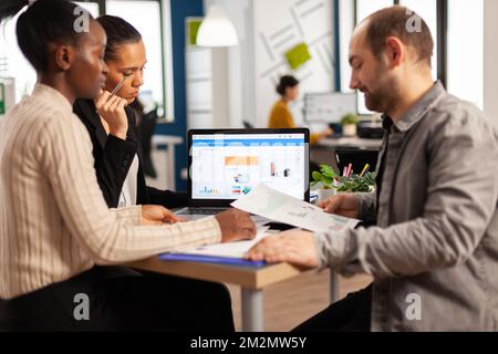 Dipendenti multiculturali impegnati e diversificati che analizzano le statistiche finanziarie annuali seduti alla scrivania di fronte a un notebook in cui sono presenti documenti in cerca di soluzioni aziendali. Team di imprenditori che lavorano in azienda Foto Stock