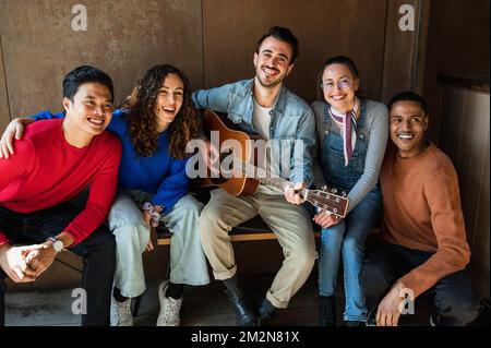 Gruppo misto di giovani amici che guardano la telecamera posando con volti felici e sorridenti mentre suonano la chitarra. Foto Stock