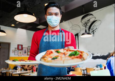 Ritratto di giovane cameriere sorridente con maschera protettiva che serve e offre piazze. Foto Stock