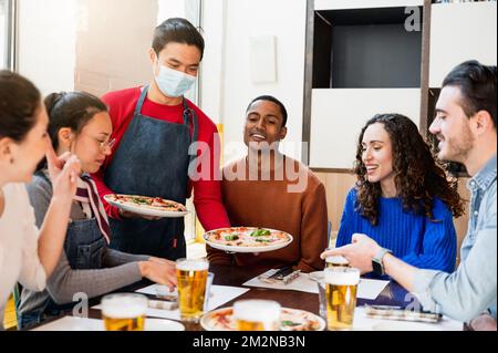 Giovane cameriere asiatico con maschera protettiva che serve pizza a un gruppo multirazziale di giovani amici seduti al tavolo del ristorante. Persone con felice s Foto Stock