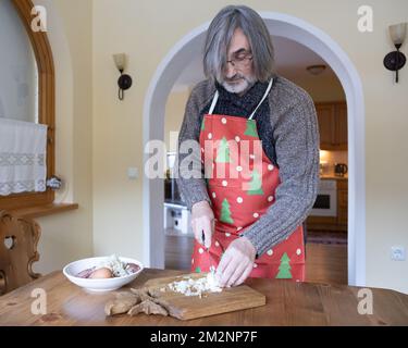 Un uomo dai capelli lunghi in un cardigan di lana e un allegro grembiule di Capodanno prepara il cibo, taglia le cipolle su una tavola di legno Foto Stock