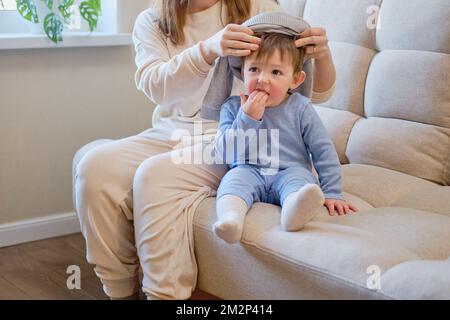 Il bambino è vestito con abiti caldi invernali per una passeggiata all'aperto. Donna madre mette i vestiti sul bambino del bambino del toddler per il tempo freddo. Bambino di un anno cinque mont Foto Stock