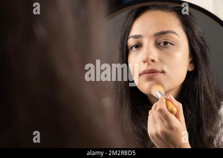 Donna che applica il trucco. Tenere e utilizzare la spazzola per fondazioni in polvere, arrossamento o liquido. Ragazza che guarda lo specchio. Bellezza, moda, stile di vita moderno. Foto Stock