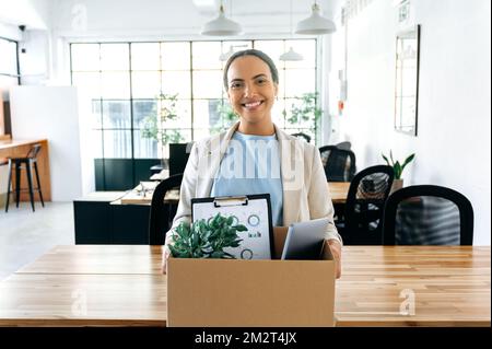 Felice latino o donna d'affari brasiliana, in abiti eleganti, in piedi nel suo nuovo ufficio di lavoro, tenendo una scatola con oggetti di lavoro in mano, ha ottenuto un nuovo lavoro, guardando la macchina fotografica, sorridendo felicemente Foto Stock