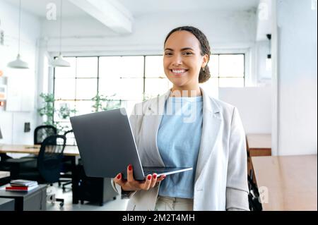 Foto di una bella donna brasiliana o latino business di successo, in abiti eleganti, seo, top manager, programmatore, si trova in un ufficio moderno, tenendo in mano un computer portatile aperto, guarda la fotocamera, sorridendo Foto Stock