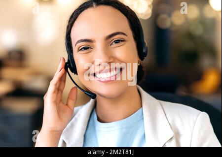 Foto ravvicinata di una giovane donna brasiliana o latina di successo, con cuffie, operatore del call center, operatore del servizio di assistenza, siede in ufficio, consulta i clienti online, guarda la fotocamera, sorridendo Foto Stock