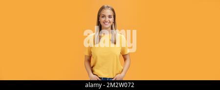 Cute chubby femmina con pelle abbronzata e bella posa dei capelli ottimista e gioioso contro sfondo arancione che tiene le mani in tasca sorridendo ampiamente a. Foto Stock