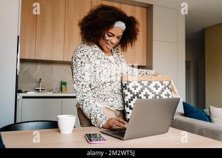 Giovane bella donna africana sorridente che tiene un cesto di loungdry lavorare con il portatile a casa Foto Stock