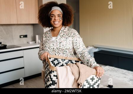 Giovane bella donna africana sorridente che tiene un cesto di loungdry e guarda la macchina fotografica a casa Foto Stock