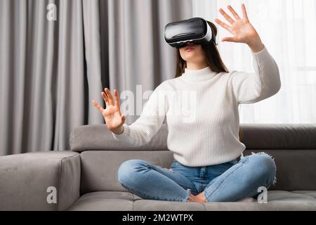 Giovane donna in occhiali vr, giocare ai videogiochi e toccare qualcosa in aria. Giocare a giochi a casa nel soggiorno Foto Stock