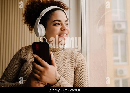 Giovane donna afro-americana felice indossando cuffie sorridenti e utilizzando il cellulare a casa Foto Stock