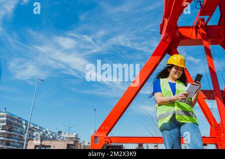 giovane ingegnere caucasico latino donna, vestito in blu con casco di sicurezza giallo e giubbotto, è in piedi all'aperto guardando il suo libro degli esercizi e mandando mes Foto Stock