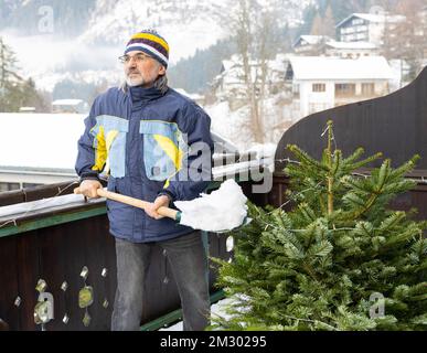 Un uomo con barba e occhiali in abbigliamento sportivo pulisce la neve con una pala sul balcone vicino all'albero di Natale Foto Stock