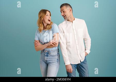 White surprised couple exclaiming and looking at each other isolated over blue background Foto Stock