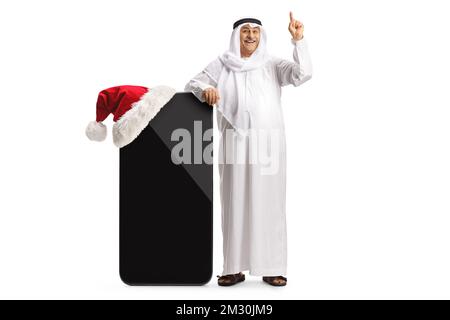 Mature arab man in a traditional robe leaning on a big mobile phone with a christmas hat and pointing isolated on white background Foto Stock