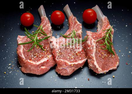 carne di agnello crudo in sella all'osso con sale, rosmarino decorato con pepe e pomodoro, vista dall'alto su sfondo nero Foto Stock