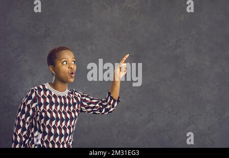 Donna afroamericana in abiti casual con sorpresa espressione facciale che punta il dito da parte. Foto Stock