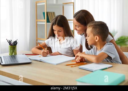 La formazione online con la mamma di un notebook aiuta i suoi figli a fare i compiti seduti alla scrivania. Foto Stock