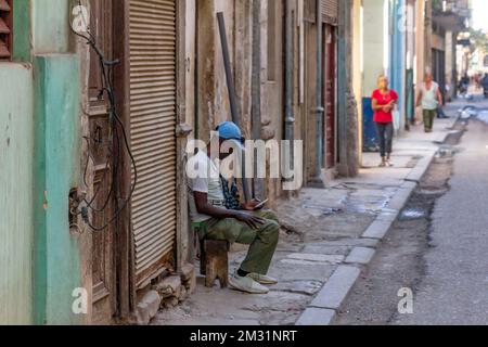 Un uomo cubano afro-caraibico siede in un marciapiede rotto e rovinabile. Gli edifici della zona sono intemperie e rovinati. Foto Stock