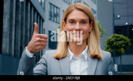 Primo piano giovane donna d'affari sorridente in tuta in piedi all'aperto contro lo sfondo di un edificio di uffici guardando la macchina fotografica mostrando il pollice in su approvazione del gesto Foto Stock