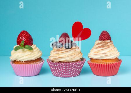 Cupcake con panna montata decorata polvere di cioccolato, fragola, lampone e mirtilli su sfondo blu, concetto di St San Valentino Foto Stock