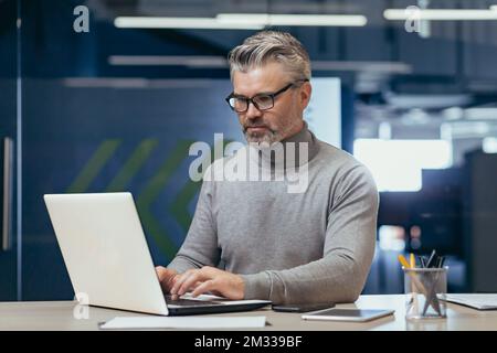 Ritratto di un uomo anziano dai capelli grigi, un uomo d'affari, direttore, fondatore, che si sta concentrando sul lavoro in ufficio alla scrivania con un computer portatile. Foto Stock