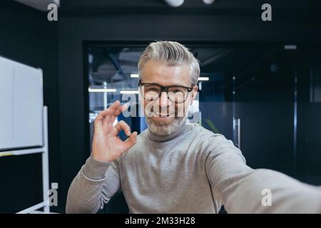 Primo piano. Senior grigio-haired businessman, freelancer prende un selfie, parla con la telecamera in una videochiamata, spara un blog. Sorride alla telecamera, mostra ok Foto Stock