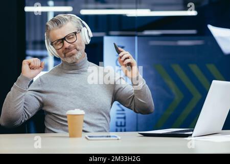 Ritratto di un uomo anziano dai capelli grigi che ascolta musica in cuffia, seduto alla scrivania in ufficio, tenendo il telefono con gli occhi chiusi, ballando, rilassato. Foto Stock
