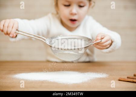 primo piano la bambina sta cucinando nella kitchen.cute capretto setaccia la farina attraverso un setaccio. Foto Stock